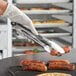 A person wearing gloves using Tablecraft locking tongs to pick up sausages over a grill.