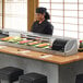 A woman standing behind a Hoshizaki refrigerated sushi display case with sushi on the counter.