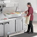 An Avantco countertop refrigerated prep rail holding food on a counter while a man in a red shirt and white gloves prepares it.