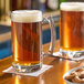 Two Anchor Hocking glass mugs of beer on a table next to a bowl of food.