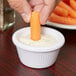 A person's fingers holding a carrot stick and dipping it into a white Carlisle ramekin.