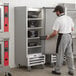 A man wearing a white shirt and grey pants opening the solid door of a Beverage-Air reach-in refrigerator.