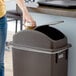 A woman uses a Lavex brown slim rectangular trash can with a dome lid to throw away a coffee cup.