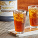A glass of Narvon Lemon Sweet Iced Tea with ice on a wood surface next to a bottle of syrup.