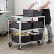 A man pushing a Regency stainless steel utility cart with black containers on the shelves.