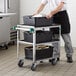 A man pushing a Regency stainless steel utility cart with black containers on the shelves.