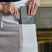 A woman putting a pen in the pocket of a white Choice full-length bistro apron.