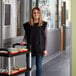 A woman wearing a black Choice cobbler apron standing in a hallway with a cart of food.