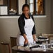 A woman wearing a white Choice poly-cotton bib apron with 2 pockets setting a table in a professional kitchen.