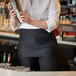 A woman wearing a black Choice standard waist apron holding a silver shaker.