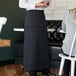 A woman wearing a black Choice full-length bistro apron with pockets standing in front of a table.