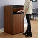 A woman standing next to a Lancaster Table & Seating walnut trash can enclosure with "THANK YOU" swing door.