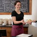 A woman wearing a burgundy standard waist apron with three pockets serving a man a cup of coffee.