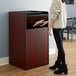 A woman standing next to a mahogany Lancaster Table & Seating waste receptacle with a "THANK YOU" swing door.