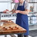 A royal blue poly-cotton standard bib apron with two pockets, worn by a person in a kitchen setting.