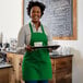 A smiling woman wearing a Choice Kelly Green poly-cotton bib apron holding a tray of coffee cups.