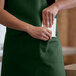 A person wearing a Choice hunter green bib apron with pockets putting a piece of paper in a pocket.