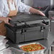 A woman in a brown apron using a CaterGator black food pan carrier to prepare food.