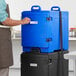 A man in an apron standing next to a blue CaterGator food pan carrier.