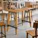 A bar height trestle table with a honey maple finish and industrial metal accents, designed for 30" x 60" table tops.