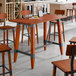 A bar height trestle table with a rustic mahogany finish and industrial metal accents, designed for 30" x 60" table tops.