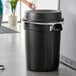 A woman throwing a white object into a black Rubbermaid BRUTE trash can.