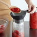 A hand using a Ball canning spoon to pour red sauce into a jar.