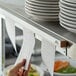 A person using a Prince Castle wall-mounted ticket holder to display a paper with writing on it over a shelf with stacks of plates.