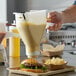 A hand using a Prince Castle mustard dispenser to put mustard on a cheeseburger.