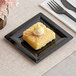 A square black WNA Comet Milan Grand plastic dessert plate with a square yellow dessert with white frosting on top.