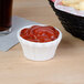 A flared white Hall China ramekin filled with ketchup on a table with a basket of french fries.