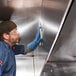 A man wearing blue gloves and goggles using 3M Stainless Steel Hood Degreaser Wipes to clean a metal surface.