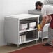 A man in a red apron putting food into a Regency stainless steel table with a metal shelf.