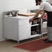 A man in a black apron opening a stainless steel cabinet on a stainless steel enclosed base table in a professional kitchen.