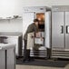 A woman in a school kitchen opening an Avantco reach-in refrigerator.