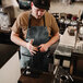 A man in a black Hardmill waxed canvas bib apron holding a cup of coffee.