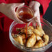 A person pouring honey garlic sauce from a small bowl onto cooked chicken wings in a glass mixing bowl.