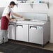 A man in a red apron and gloves using an Avantco 3 door refrigerated sandwich prep table to prepare food in a commercial kitchen.