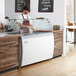 An Avantco flat glass sneeze guard on a counter with a man in a white apron.