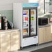 A white sliding glass door merchandiser refrigerator with LED lighting, stocked with beverages and placed between countertops in a commercial setting.