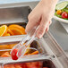 A hand holding a pair of Cambro plastic tongs with ketchup dipping a tomato.