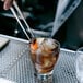 Arcoroc by Chris Adams garnish tongs being used to garnish a glass of brown liquid with ice and an orange peel.