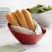 A red slanted melamine bowl filled with bread sticks on a table with a salad.