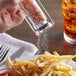 A hand using a Regal pink Himalayan salt shaker to sprinkle salt over a plate of french fries.