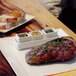 A piece of steak with green sauce in a white rectangular ramekin on a plate.