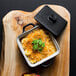 A black square stoneware casserole dish with a lid on a wood cutting board.