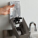 A hand using a Rubbermaid metallic gray manual spray soap dispenser to fill a plastic bag with liquid soap.