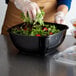 A woman in gloves preparing a salad in a Fineline black plastic bowl.