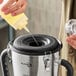 A stainless steel commercial food blender with a black lid, being filled with liquid from a squeeze bottle.