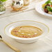 A bone or ivory plastic bowl with gold bands, filled with a creamy soup and garnished with herbs, placed on a table.